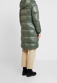 Grüner gesteppter Mantel mit glänzendem Finish, langer Länge, angenähter Kapuze, elastischen Bündchen und tailliertem Schnitt. Kombiniert mit beigen Hosen und schwarzen Stiefeln.