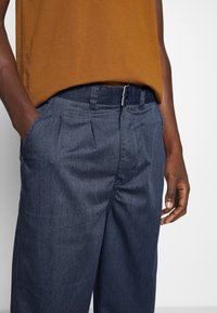 Mörkblå plisserade byxor i texturerat tyg, med sidofickor och en midja med silverfärgad knappstängning.