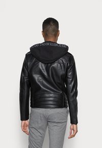 Giubbotto in pelle nera con texture a righe, dotato di cappuccio nero con marchio bianco lungo il bordo. Modello che indossa pantaloni grigi.