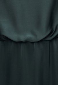 Σκούρο πράσινο λείο ύφασμα με απαλά πτυχώσεις και ραφή που συλλέγεται, που δείχνει υφή και ελαφριά γυαλάδα.