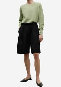 Jasnozielony dzianinowy sweter, czarne plisowane szorty i płaskie czarne buty. Sweter ma długie rękawy; szorty sięgają powyżej kolana, o kroju dopasowanym.