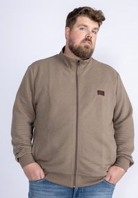 Hnědá zipová bunda s žebrovanou texturou a malou červenou nášivkou na levé hrudi. Má vysoký límec a elastické manžety.