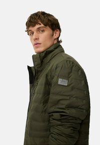 Gepolsterte olivgrüne Jacke mit hohem Kragen, seitlichen Reißverschlusstaschen und grauem Logo-Patch am Ärmel. Sie verfügt über ein schlankes, stromlinienförmiges Design.