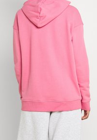 En person som bär en ljusrosa hoodie och ljusgråa sweatpants, står med ryggen mot kameran.