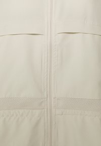 Ljusbeige zip-up jacka med slät tyg, som har två framfickor och ett perforerat texturdetalj i den nedre delen.