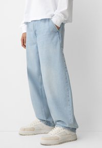 Svetlo modri široki traper hlače z zbledelim zaključkom, ki imajo stranske žepke in so v kombinaciji z belimi supergami. Material se zdi denim z mehko teksturo.