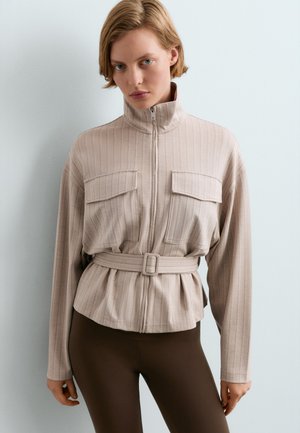 Femme portant une veste beige zippée avec des poches poitrine et une ceinture, associée à un pantalon marron foncé, debout contre un mur clair uni.
