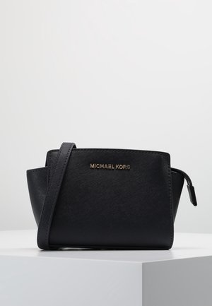 Zwarte Michael Kors crossbodytas met een gestructureerd oppervlak, gouden logo en verstelbare band op een wit voetstuk tegen een effen achtergrond.