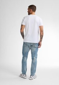 Camiseta de algodón blanca con escote redondo, llevada con jeans de mezclilla azul claro. Tatuajes visibles en el brazo, de pie contra un fondo neutro.