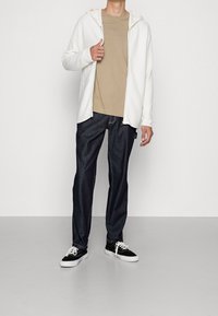 White zip-up hoodie with textured fabric, beige t-shirt, dark denim jeans, and black sneakers with white soles. Simple contemporary design.