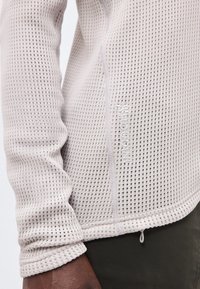 Κοντινό σε πρόσωπο που φοράει ανοιχτό μπεζ μακρυμάνικη μπλούζα με υφή και σχέδιο πλέξης βάφλας, με ορατές λεπτομέρειες ραφής στα πλαϊνά.