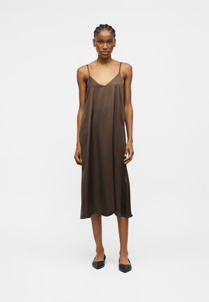 Frau steht und trägt ein ärmelloses braunes Midi-Negligékleid mit dünnen Trägern und schwarze spitze Ballerinas vor einem schlichten weißen Hintergrund.