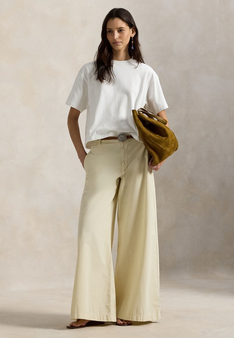 T-shirt blanc en coton à manches courtes, associé à un pantalon beige à jambes larges. La tenue est complétée par un sac à main en daim marron et une boucle de ceinture décorative.