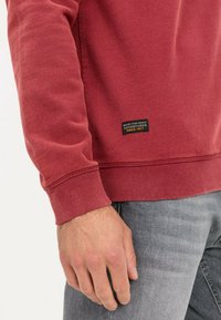 Sweat-shirt rouge avec poignets et ourlet côtelés, tissu texturé, avec une étiquette noire avec du texte sur le côté inférieur. Jean gris visible en dessous.