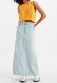 Nainen, joka on pukeutunut hihattomaan oranssiin neulecrop-toppiin, vaaleansiniset korkeavyötäröiset farkku-maxihameeseen ja valkoisiin lenkkareihin mustilla yksityiskohdilla.