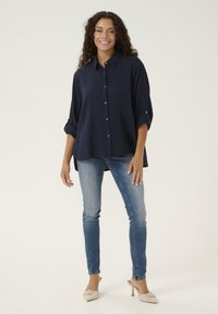 Camicia blu navy con bottoni e tessuto a trama, maniche arrotolate e orlo curvo, abbinata a jeans blu chiaro e tacchi beige.