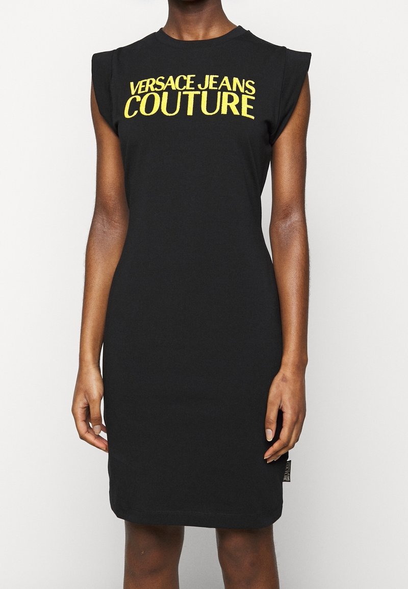 Robe noire sans manches avec le texte jaune « Versace Jeans Couture » sur la poitrine portée par une personne debout devant un fond uni.