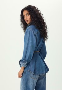 Camicia di jeans in blu medio, caratterizzata da un design con legatura posteriore, maniche lunghe e vestibilità rilassata con orlo arrotondato e dettagli di cucitura in vista.
