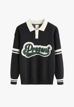 Pull en tricot noir avec un col blanc, arborant un logo "Cadeau" en fausse fourrure verte et des accents rayés blancs sur les manches.
