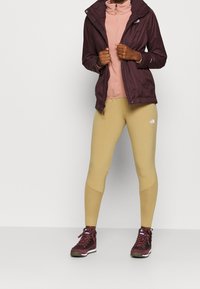 Bordeaux lichtgewicht jack over een roze fleece, gecombineerd met beige leggings en bordeauxkleurige wandelschoenen. De outfit heeft verschillende texturen en kleuren.