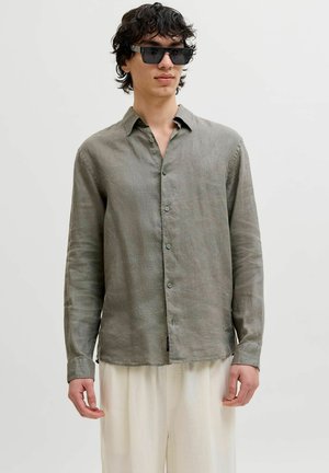 Ung mann iført svarte firkantede solbriller, løs grå skjorte med knapper og lyse bukser, stående mot en ensfarget bakgrunn.