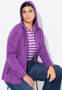 Lila fleece zip-hoodie med en strukturerad yta, justerbar huva och sidofickor, bärs över en randig lila och vit t-shirt.