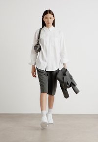 White long-sleeve button-up shirt, gray tailored shorts, white sneakers, and carrying a gray blazer and small bag. Smooth textures, neutral colors.