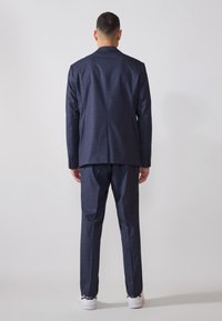 Costume bleu marine pour homme avec un motif à carreaux, coupe ajustée, revers cranté et pantalon assorti. Baskets blanches visibles en bas.