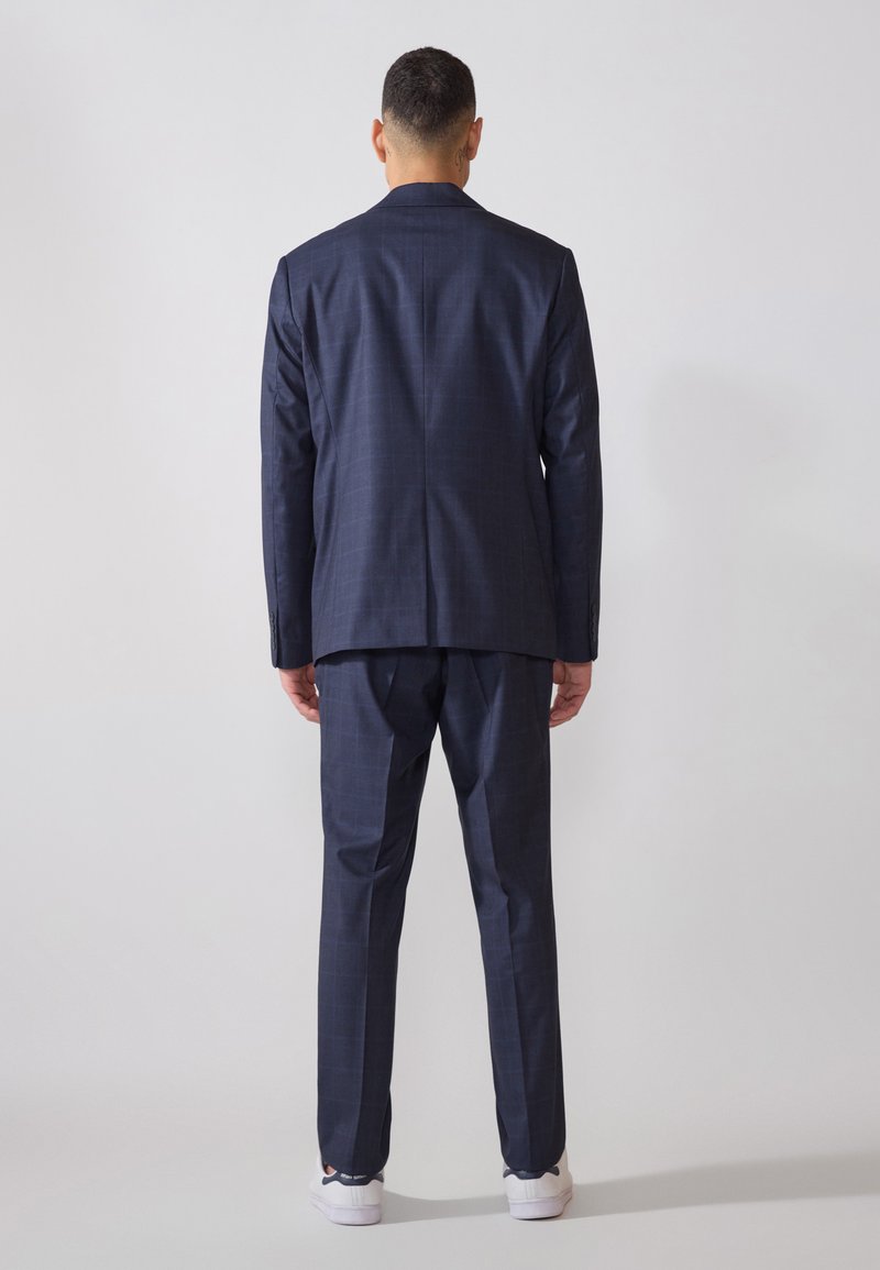 Costume bleu marine pour homme avec un motif à carreaux, coupe ajustée, revers cranté et pantalon assorti. Baskets blanches visibles en bas.