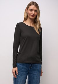Junge Frau mit langen blonden Haaren, die ein schwarzes Langarmshirt und blaue Jeans trägt, vor einem schlichten hellen Hintergrund stehend.