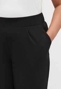 Black trousers with a smooth texture and an elastic waistband; features side pockets and subtle pleating for added detail.