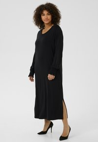 Vestido negro de manga larga hecho de una tela suave, con escote en forma de barco, puños fruncidos y aberturas laterales. Combinado con tacones altos negros.