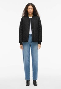 Črno prešito bomber jakno z rebrastimi manšetami, sprednjim zadrgo in stranskimi žepi, kombinirano s svetlo modrimi kavbojkami z visokim pasom in črnimi gležnjnimi škornji.