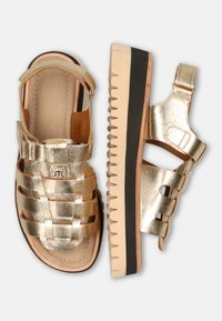 Metallieke gouden sandalen met geweven banden, verstelbare enkelgesp en een gestructureerde beige loopzool met een zwarte rand voor grip.