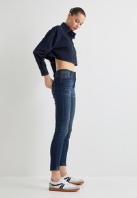 Camisa de botones azul marino, jeans oscuros ajustados con dobladillo deshilachado, y zapatillas blancas con acentos negros y suelas marrón.
