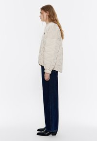 Giubbotto imbottito beige con motivo a zigzag, polsini elasticizzati e orlo laterale. Abbinato a jeans di denim scuri a vita alta e stivali neri alla caviglia.