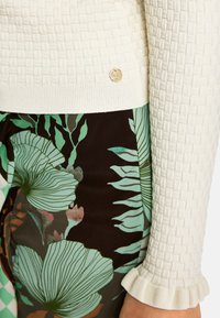 Gestrickter cremefarbener Pullover mit einem strukturierten, gitterartigen Muster; verfügt über einen schlichten Saum und ein rundes goldenes Logo-Detail nahe dem Saum. Kombiniert mit floral bedruckten dunklen Hosen in grünen und braunen Farbtönen.
