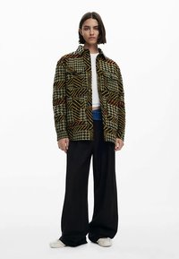 Multicolored patterned jacket with houndstooth and stripes, featuring two chest pockets, button closure, and a relaxed fit, worn over a white top.