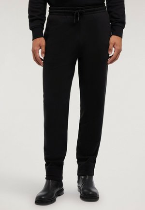 Hombre con pantalones de chándal negros con cordón y botines de cuero negros, de pie frente a un fondo liso.