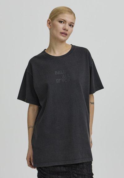 Szőke nő hátrafésült hajjal, kopott fekete, túlméretezett pólót visel, amelyen halvány szöveg látható, egyszerű szürke háttér előtt áll.