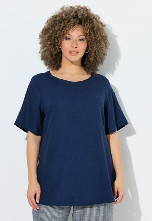 Kobieta z kręconymi włosami, nosząca kolczyki-koła, luźny granatowy top z krótkim rękawem oraz szare spodnie w cienkie prążki, stoi na białym tle.