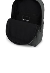 Rucsac din țesătură neagră cu o textură netedă, buzunar frontal cu fermoar și un patch cu logo proeminent. Dispune de un compartiment principal spațios.