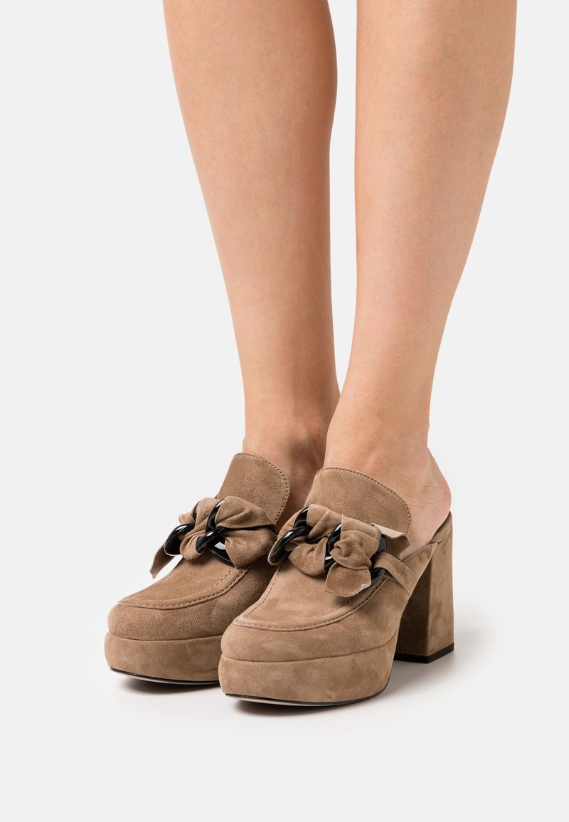 Mules compensées en daim marron avec une grande chaîne noire et un détail nœud, portées pieds nus sur fond blanc.