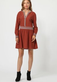 Vestido de color óxido con mangas largas, escote en V y bordados estampados a lo largo del escote y los puños. Falda acampanada con una textura suave.