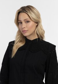 Zwarte blouse met een gegolfde kraag, gestructureerd lijfje en geborduurde kantmouwen, met een knoopsluiting aan de voorkant en subtiele verticale strepen.