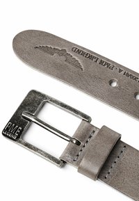 Ceinture en cuir gris avec boucle en métal gravée "PME Legend" et motif d'aile en relief près des trous perforés sur fond blanc.