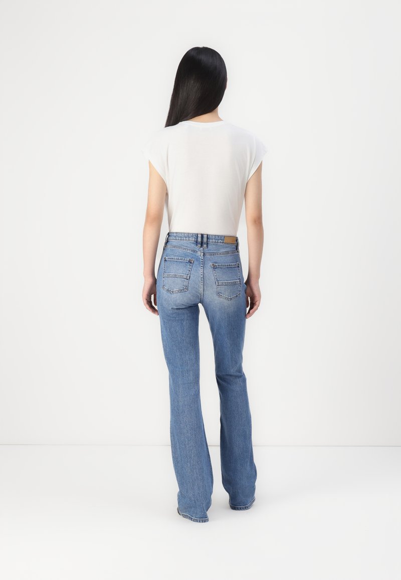 Weißes Oberteil mit kurzen Kapuzeärmeln, kombiniert mit hellblauen, ausgestellten Jeans aus Denim, die zwei Gesäßtaschen und ein Taillenschild haben. Glatte Textur, lässige Passform.