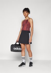 Débardeur à motif rouge, jupe de sport noire avec des rayures blanches, chaussures de sport noires, chaussettes blanches avec des rayures noires, portant un sac noir Adidas.