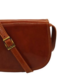 Sac en cuir marron à bandoulière avec un rabat courbé, une sangle ajustable et une boucle en laiton. Les accents cousus rehaussent le design.