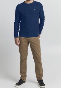 Granatowy sweter z dzianiny z teksturowanym panelem na klatce piersiowej, okrągłym dekoltem i ściągaczami na rękawach, w zestawie z khaki spodniami i czarnymi sneakersami.